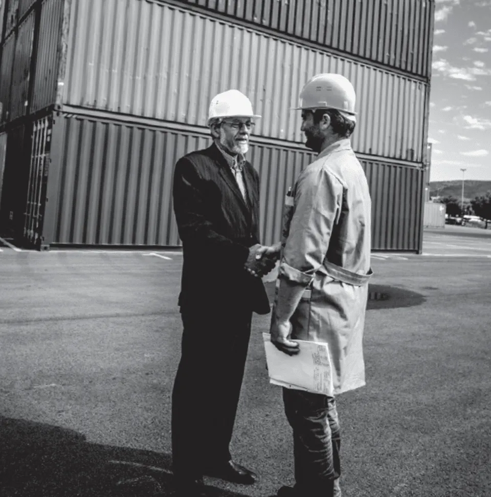 Two terminal workers in front of stacked shipping containers shaking hands